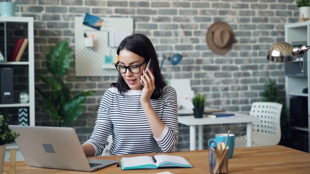 Femme au téléphone devant un ordinateur portable et un carnet ouvert